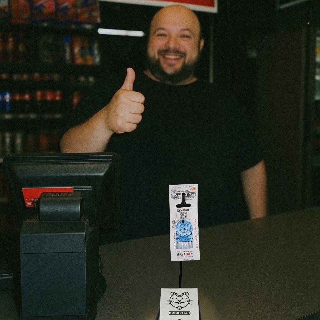 Lächelnder Shopbesitzer hinter dem Tresen gibt Daumen nach oben neben einem LUCKY TO HAVE® Verkaufsständer mit Logo und Produkt.
