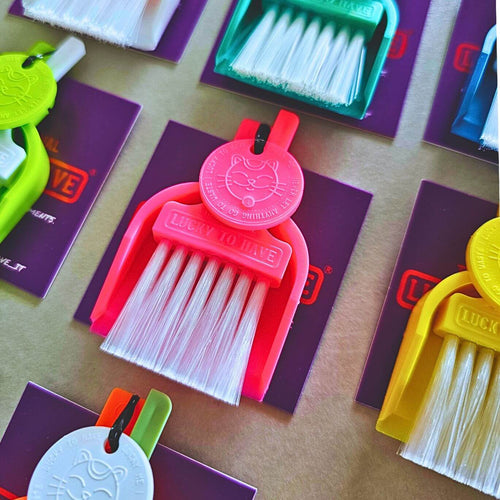 Hot pink LUCKY TO HAVE®, the world’s smallest broom set, with white bristles and a matching Lucky Coin, surrounded by colourful sets on purple product tags.