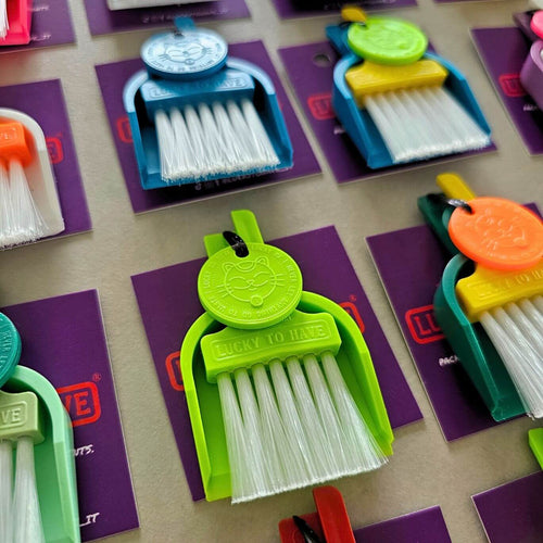 A grid of LUCKY TO HAVE® mini dustpan-and-brush sets in assorted colors — blue, lime green, teal, coral, and orange — each clipped to a purple backing card. A bright green set is in sharp focus in the foreground, showing its round cat-face tag charm and "LUCKY TO HAVE" embossed on the dustpan, with white bristles fanned out below.
