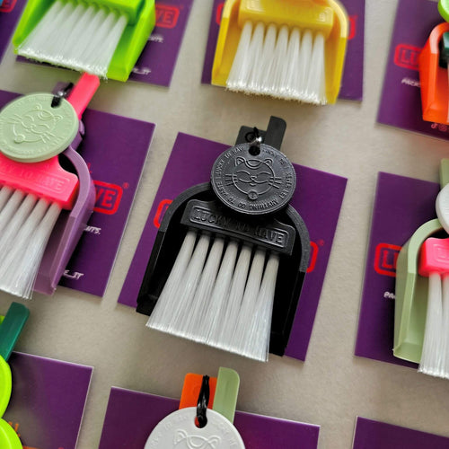 Black LUCKY TO HAVE® mini broom set for powders and fine crumbs, with white bristles and a matching black Lucky Coin, surrounded by colourful sets on purple tags.