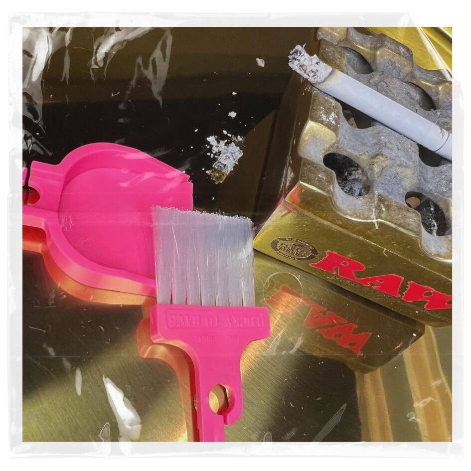 LUCKY TO HAVE® pink mini dustpan and broom placed beside a RAW ashtray, sweeping fine cigarette ash from a golden tray for precise cleanup.