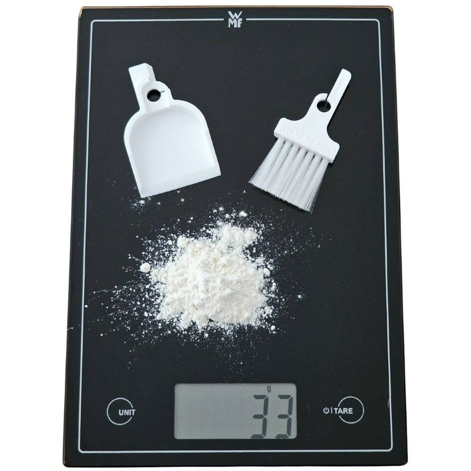 LUCKY TO HAVE® Lucky Blanco mini dustpan and broom placed on a WMF digital kitchen scale next to fine white powder, enabling precise measuring and clean handling of delicate substances - fine things matter.