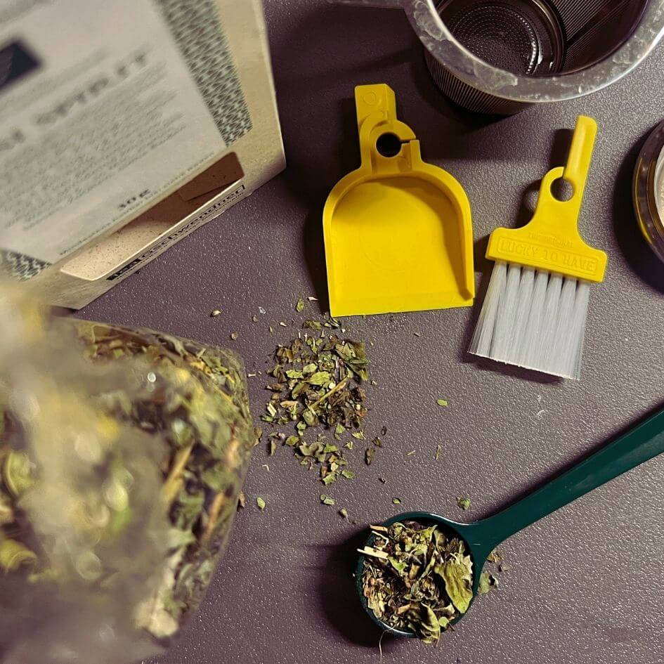 LUCKY TO HAVE® Banana yellow smallest broom and dustpan set next to loose tea leaves, showing dried tea herbs scattered on a tabletop with spoon and tea packaging in view.