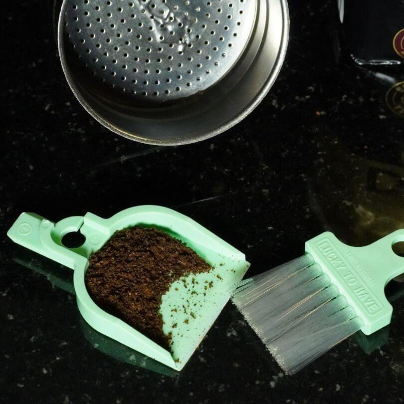 Mintfarbenes Kehrset entfernt verschüttetes Kaffeepulver neben einem Espressomaschinen-Sieb.