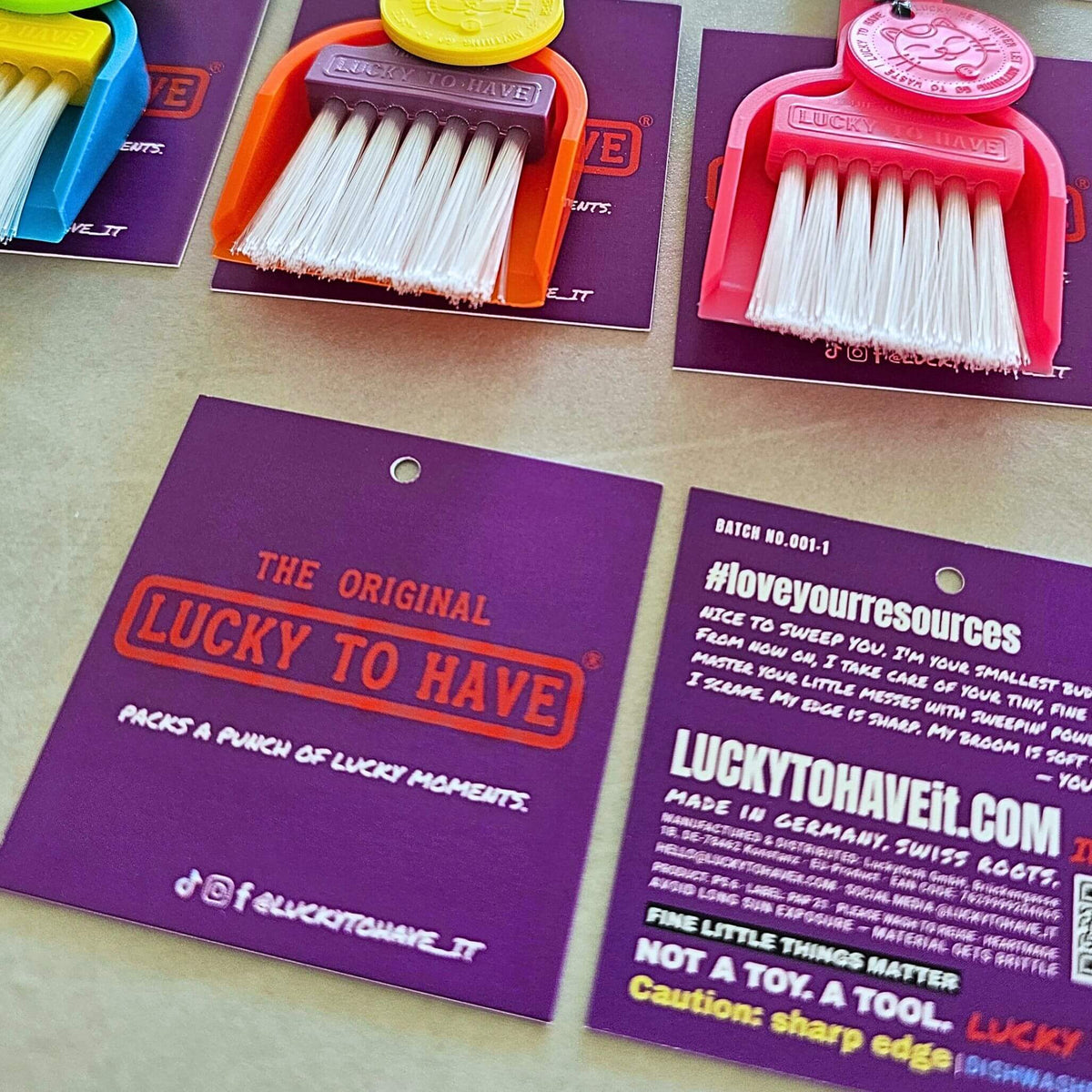 Colourful LUCKY TO HAVE® mini broom sets with white bristles and Lucky Coins on purple product tags, with tag fronts and backs displayed alongside.
