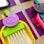Close-up of a LUCKY TO HAVE® mini broom set with a lime green brush, yellow dustpan and purple Lucky Coin, for sweeping powders and fine crumbs.