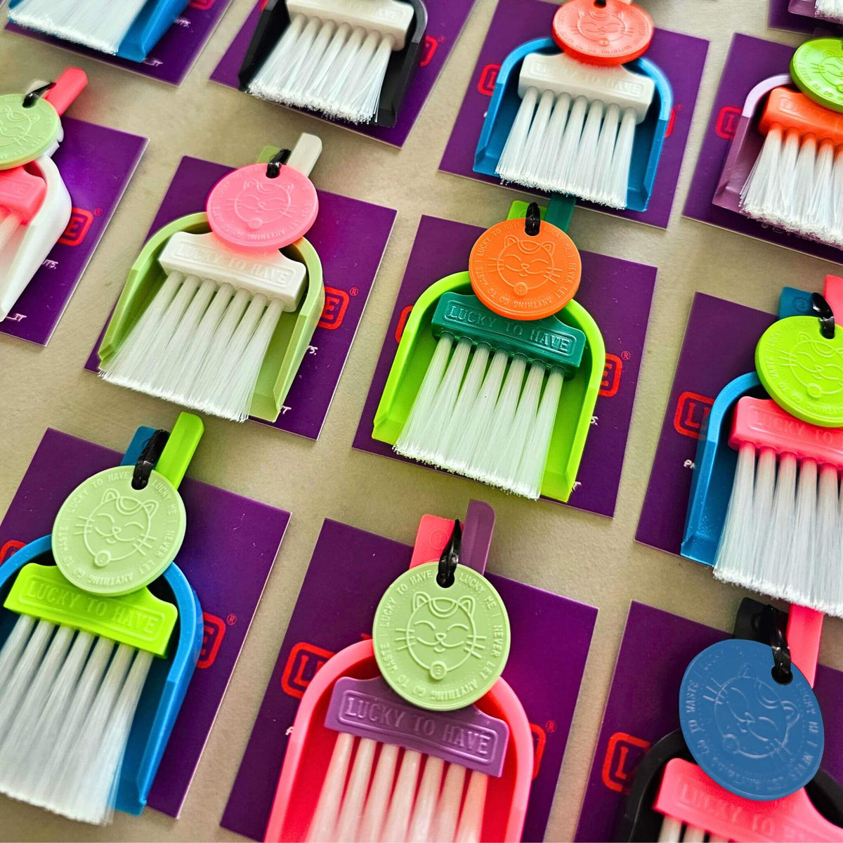 Rows of LUCKY TO HAVE®, the world’s smallest broom set, in assorted colours, including a dark green brush with a lime dustpan and orange Lucky Coin.