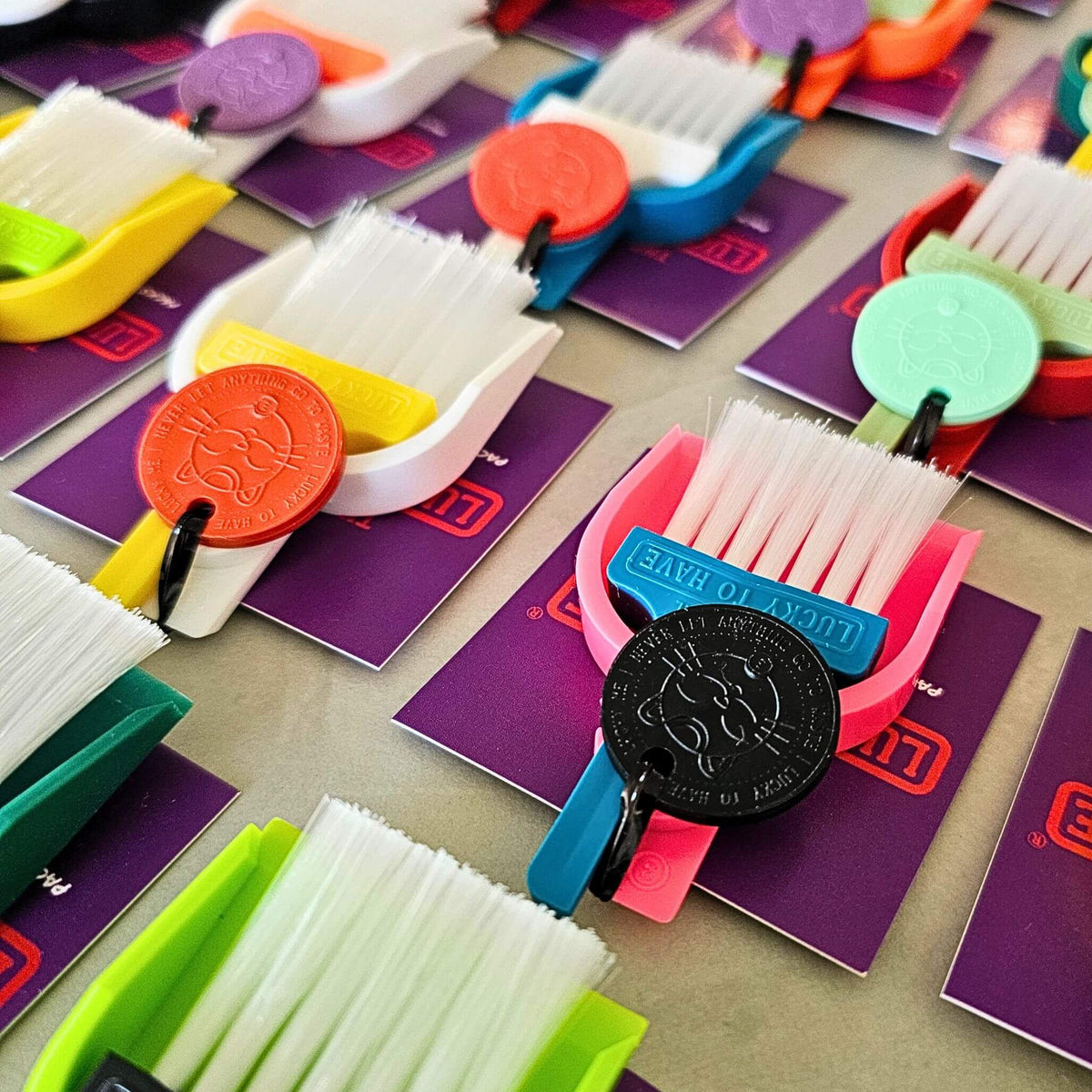 Colourful LUCKY TO HAVE® mini broom sets arranged on purple tags, featuring a blue brush in a pink dustpan with a black Lucky Coin.
